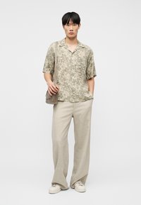 Νεαρός άνδρας στέκεται, φοράει μπεζ κοντομάνικο πουκάμισο με λουλουδάτο σχέδιο, ανοιχτό μπεζ παντελόνια με φαρδιά πόδια, λευκά αθλητικά παπούτσια και κρατά μία ώμου τσάντα σε απόχρωση ταυ.