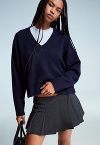Ženska z dolgimi pletenicami, oblečena v mornarsko modro pulover s V-izrezom nad belim zgornjim delom, s temno prugasto nabrano krilo, drži črno torbico.
