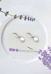 Silverfärgade krokörhängen med klara, facetterade ovala stenar, placerade på en vit tallrik omgiven av lavendelkvistar.