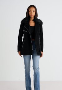 Čierna bunda s umelou kožušinou na límci, s vreckami na zips a asymetrickým dizajnom zipsu; spárovaná so svetlomodrými, mierne rozšírenými džínsami.