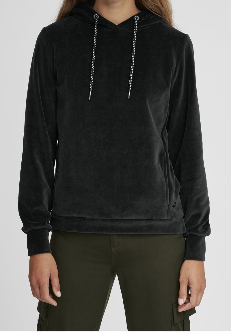 Personne portant un hoodie en velours noir avec des cordons blancs et un pantalon cargo vert olive, debout devant un fond gris uni.