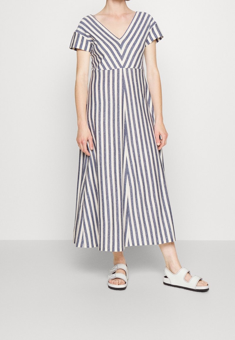 Femme portant une robe rayée avec un col en V et des manches courtes, associée à des sandales blanches ouvertes à l'avant, debout devant un fond uni.