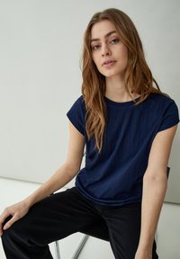 Camiseta de manga corta azul marino con cuello redondo; hecha de una tela texturizada. Lleva pantalones negros, modelo sentado en una silla.