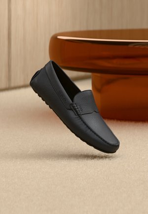 Mocassin en cuir noir à surface texturée, flottant légèrement au-dessus d'un tapis beige, mobilier moderne arrondi marron en arrière-plan flou.