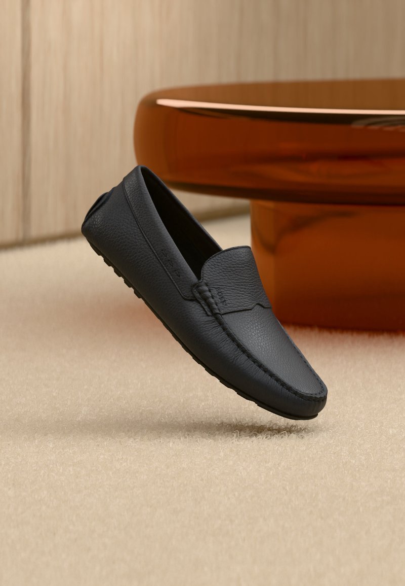 Mocassin en cuir noir à surface texturée, flottant légèrement au-dessus d'un tapis beige, mobilier moderne arrondi marron en arrière-plan flou.