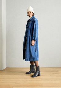 Trench coat de mezclilla en azul con diseño de doble botonadura, grandes solapas y bolsillos laterales. Botines negros con tacones en bloque y textura suave.