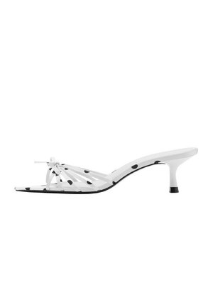 Sandalia de tacón tipo gatito blanca con lunares negros, tiras delgadas y un pequeño lazo en la parte frontal, vista lateral sobre fondo blanco.