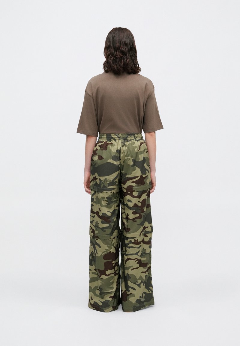 Person mit schulterlangen dunklen Haaren, die von hinten schaut, trägt ein braunes kurzärmliges Hemd und eine grüne camouflagefarbene weite Hose.
