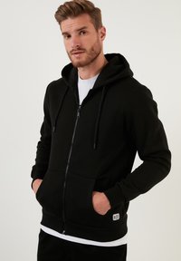 Crna dukserica na zip s prednjim džepom, kapuljačom s vezicom, rebrastim manžetama i donjim rubom; sadrži malu oznaku brenda na lijevom džepu.