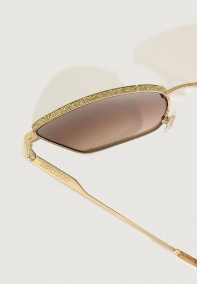 Óculos de sol em tom dourado com uma borda superior acentuada por glitter, lentes castanhas em degradê e um design elegante e curvado. Apresenta uma haste de marca.