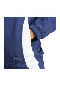 Giacca sportiva blu navale con accenti bianchi sui lati, dotata di polsi elasticizzati e di un piccolo logo "AEROREADY" vicino all'orlo. Tessuto liscio e leggero.