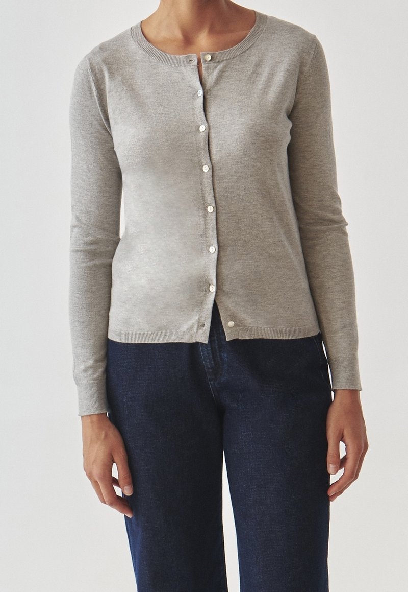 Personne portant un cardigan gris clair à boutons et manches longues, et un pantalon bleu foncé, sur un fond uni.