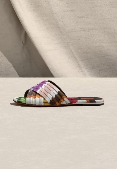 Sandale plate à semelle plate avec des brides tissées multicolores dans des tons de violet, rose, blanc, orange, vert et marron sur une surface neutre.