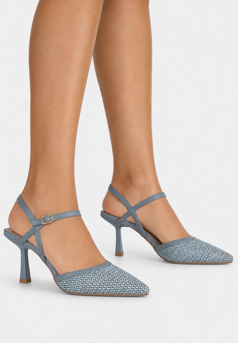 Benen met blauwe geweven slingbackhakken met puntige neus, dunne enkelbandjes en uitlopende stilettohakken op een effen achtergrond.