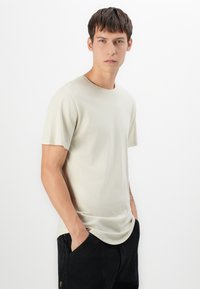 Camiseta beige claro de tela suave con escote redondo y mangas cortas, combinada con pantalones negros. Textura lisa, ajuste relajado.