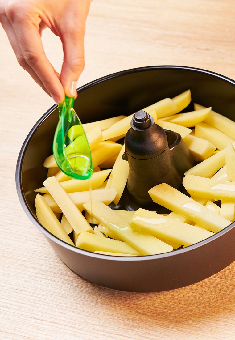 Schwarze Schüssel, gefüllt mit gelben Kartoffelstreifen; ein grüner Öldispenser träufelt Öl auf die Kartoffeln, die um einen zentralen Kegel angeordnet sind.