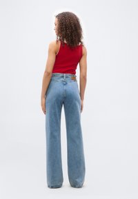 Γυναίκα με σγουρά μαλλιά στέκεται με την πλάτη, φοράει κόκκινο αμάνικο τοπ και ψηλόμεσο φαρδύ μπλε τζιν, μπροστά σε απλό λευκό φόντο.