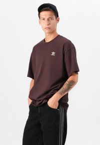 Tmavohnedé bavlnené tričko s krátkymi rukávmi a okrúhlym výstrihom. Na hrudi sa nachádza malé biele logo Adidas. Nosené s čiernymi džínsami.