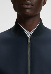 Navy bomber jacket with a textured surface, ribbed collar, and silver zipper. Features a striped shirt collar peeking from underneath.