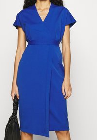 Blå omslagsklänning gjord av ett mjukt tyg, med korta ärmar, en åtsittande midja och en slits på sidan. Håller en svart väska.