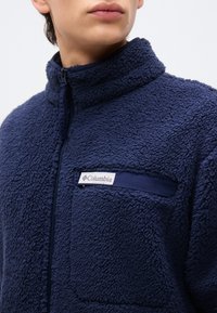 Κοντινό πλάνο ενός ατόμου που φοράει μπουφάν από φλις σε σκούρο μπλε χρώμα με φερμουάρ και τσέπη στο στήθος με φερμουάρ, με την ετικέτα "Columbia".