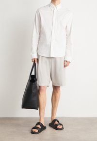 Camicia bianca con bottoni e un piccolo logo, pantaloni corti grigi chiari, sandali neri con fibbie e una grande borsa nera a tracolla. Sfondo semplice.