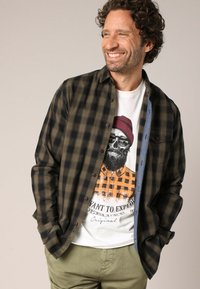 Glimlachende man met krullend haar, die een open donker groene en zwarte geruite overhemd draagt over een witte T-shirt met een afbeelding van een schedel die een beanie en zonnebril draagt.
