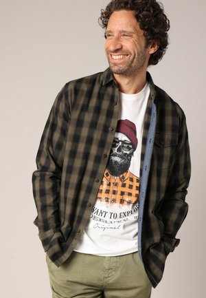 Glimlachende man met krullend haar, die een open donker groene en zwarte geruite overhemd draagt over een witte T-shirt met een afbeelding van een schedel die een beanie en zonnebril draagt.