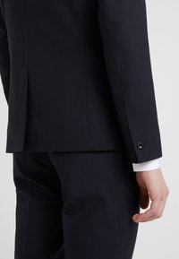 Marineblauer maßgeschneiderter Anzug mit glatter Textur, mit einem Schlitzausschnitt auf der Rückseite und Knopfdetails an den Ärmeln. Weisses Hemd darunter sichtbar.