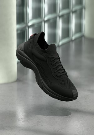 Baskets de sport noires avec tige en tricot, lacets et semelle coussinée flottant au-dessus d'un sol en béton poli près de fenêtres en verre dépoli.