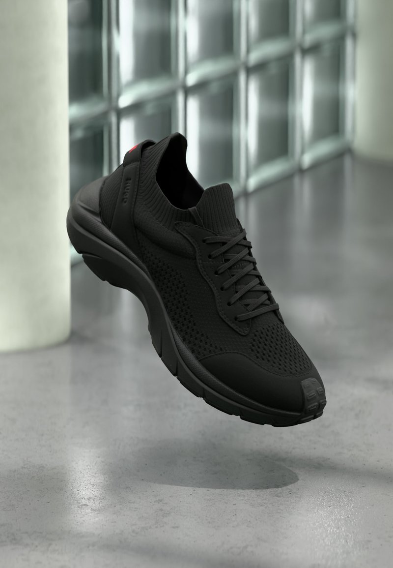 Baskets de sport noires avec tige en tricot, lacets et semelle coussinée flottant au-dessus d'un sol en béton poli près de fenêtres en verre dépoli.