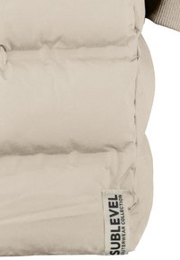 Beige gepolsterte Weste aus weichem Stoff mit einem Etikett, das "SUBLEVEL OUTERWEAR COLLECTION" liest. Sie hat eine gesteppten Textur und einen runden Kragen.