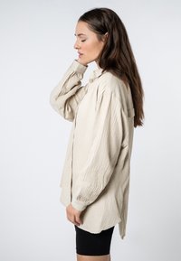Beige, textured, long-sleeve shirt with a button-down collar, slightly loose fit, and a rounded hem. Worn over black shorts.
