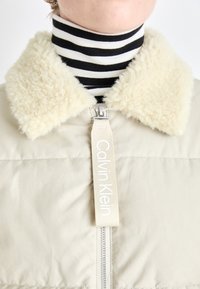 Veste en duvet beige avec un col en fausse fourrure, dotée d'une fermeture éclair à l'avant et d'une étiquette logo. La couche intérieure montre un col roulé rayé noir et blanc.