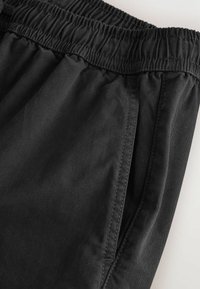 Pantalon à taille élastique noire avec poche latérale visible et détails de couture sur un tissu lisse.