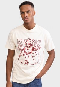 T-shirt de algodão creme com estampa vermelha do Pai Natal e o texto "Nada para ti." Mangas curtas e design clássico com gola redonda.