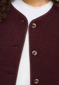 Burgundia fliisvest, tekstuuriga pinnaga, ümar kaelus, kontrastset värvi nuppe ning sileda viimistlusega ääred.
