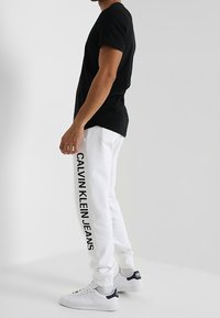 Czarny t-shirt z krótkim rękawem w połączeniu z białymi spodniami dresowymi z pionowym logo "CALVIN KLEIN JEANS". Buty sportowe są głównie białe z czarnymi akcentami.