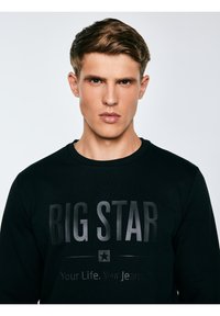 Jauns vīrietis ar īsiem brūniem matiem, tērpies melnā sporta krekliņā ar tekstu "BIG STAR Your Life. Your Jeans.", neitrāla fona priekšā.