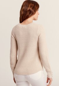 Pull en tricot beige avec un motif texturé, col rond et accents de boutons sur les manches, conçu pour un look ajusté à la taille.