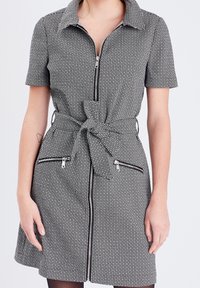 Robe à motifs noir et blanc avec manches courtes, col et fermeture éclair frontale, dotée d'une ceinture nouée et de deux poches zippées.