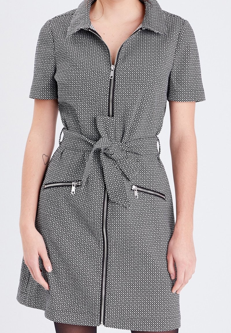 Robe à motifs noir et blanc avec manches courtes, col et fermeture éclair frontale, dotée d'une ceinture nouée et de deux poches zippées.