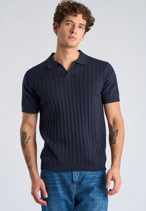 Młody mężczyzna o kręconych włosach, ubrany w granatową prążkowaną koszulkę polo z krótkim rękawem i niebieskie jeansy, stojący na tle jednolitego jasnego tła.