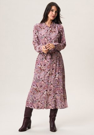 Vrouw draagt een midi-jurk met lange mouwen en knopen, bedrukt met een paisley-patroon in paarse tinten, voorzien van een taillekoord en donkere bruine laarzen met een middelhoog hakje.