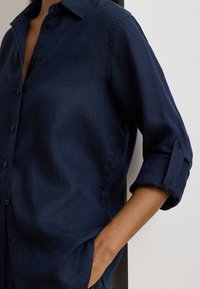 Tamsiai mėlyna mygtukais užsegama marškinėliai iš lengvos medžiagos, su apykakle, suvyniotomis rankovėmis ir laisvo silueto su šiek tiek blizgesio.