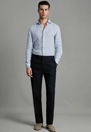 Cămașă albastră deschis din in, cu butoni și mâneci lungi, asociată cu pantaloni întunecați și pantofi slip-on de culoare maro deschis, amplasată pe un fundal gri.