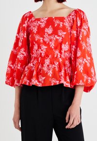 Czerwona bluzka w kwiaty ma kwadratowy dekolt, bufiaste rękawy oraz marszczony dół w kształcie peplum. Tkanina ma teksturowany, lekki wygląd.