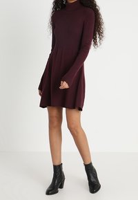 Burgundin pitkähihaine mekko, jossa on ribbed-kappale, A-linjainen hame ja kaulus. Yhdistetty mustien korkokenkäisten nilkkasaappaiden kanssa. Sujuva rakenne.