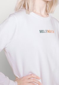 Sudadera blanca con cuello redondo, que presenta un bordado multicolor de "HILFIGER" en verde y naranja. Textura suave y corte relajado.