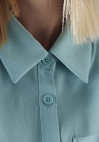 Camisa azul clara con una textura suave, que presenta un cuello puntiagudo, dos botones y un bolsillo en el pecho. Vista en primer plano que captura los detalles.